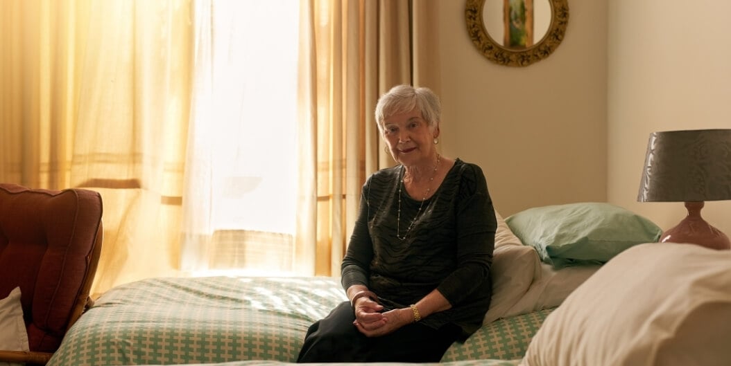 Elderly Woman Sitting on Bed Smiling at Camera in Independent Living